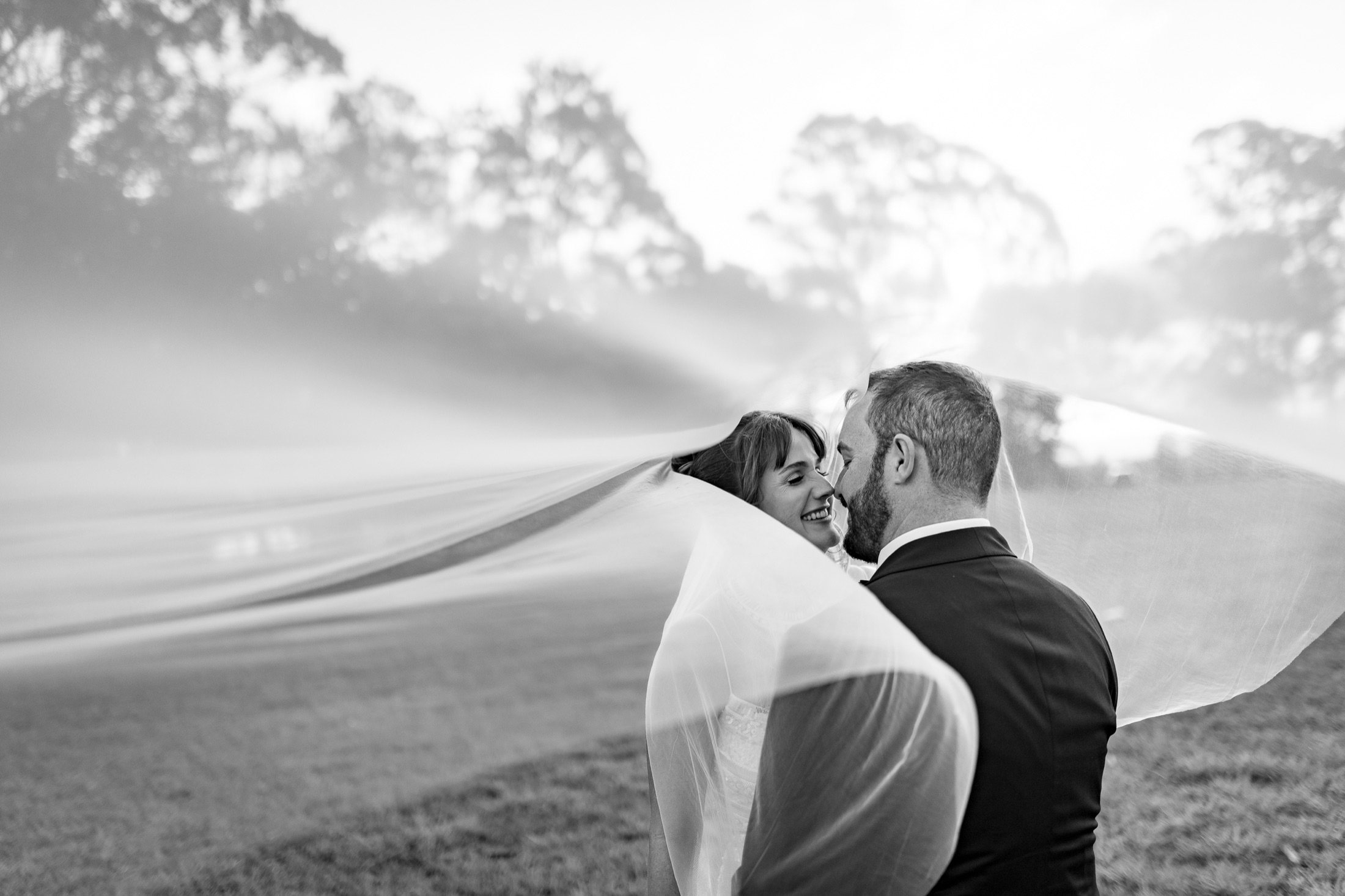 Black and white photo of a bride and groom nose to nose under a windswept veil · Jamie Hall, Definitive Imaging